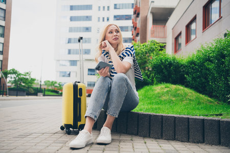 Charming young woman sitting with yellow suitcase, enjoying leisurely moments in urban park during daylightの写真素材