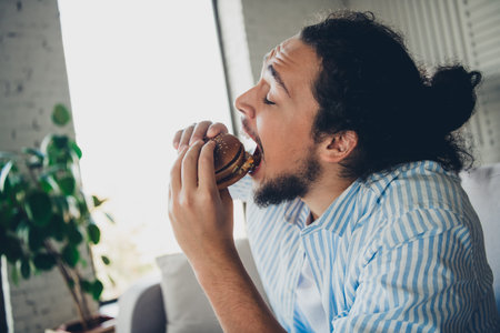 Young man enjoying a burger in a cozy home environment during daytime