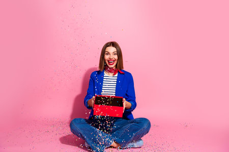 Cheerful woman opening a gift box with confetti on a pink background, wearing a blue jacket and red scarf, celebrating a festive moment.の写真素材