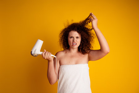 Confused young woman with curly hair holding hair dryer in towel against yellow backgroundの写真素材