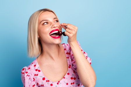 Photo of nice lovely girl wear trendy pink print clothes eat ripe cherries look empty space isolated on blue color backgroundの写真素材