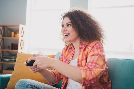Young woman with curly hair enjoying a video game on a sofa in a bright living room, expressing joy and relaxationの写真素材