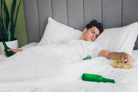 Young man lounging in a cozy bed surrounded by snack leftovers and bottles in a brightly lit bedroomの写真素材