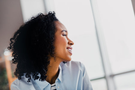 Young woman smiling brightly near a large window in a cozy living room on a sunny day, embracing relaxation and a positive lifestyle.の写真素材
