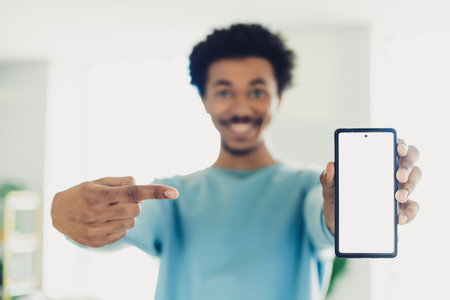 Young man in casual blue sweatshirt smiling and presenting smartphone with blank screen, indicating display while standing indoorsの写真素材