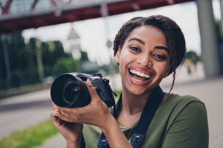 Joyful young woman holding a camera outdoors, wearing a stylish casual outfit, capturing the vibrant urban scenery on a sunny dayの写真素材