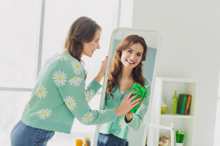Young woman in a light floral cardigan holding mirror and smiling during daylight at her cozy home interiorの写真素材