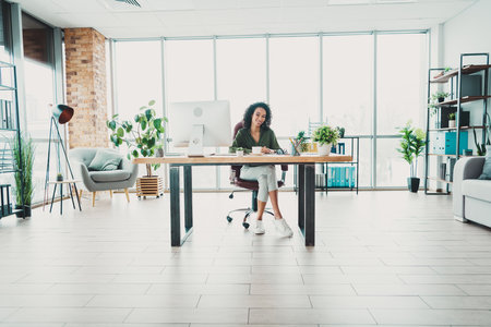 Modern open-plan office with a joyful young professional woman at a desk surrounded by greenery and illuminated by natural lightの写真素材