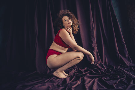 Young woman in elegant red lingerie with curly hair posing confidently against a dark purple studio backgroundの写真素材