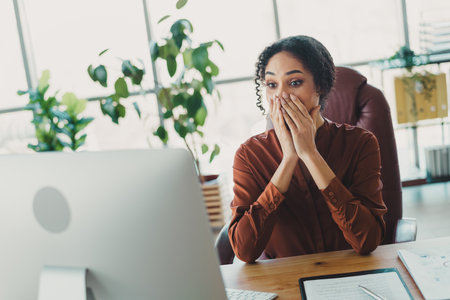 Young businesswoman in modern office surprised by news on her computer, showcasing authentic emotions and a professional workspace.の写真素材