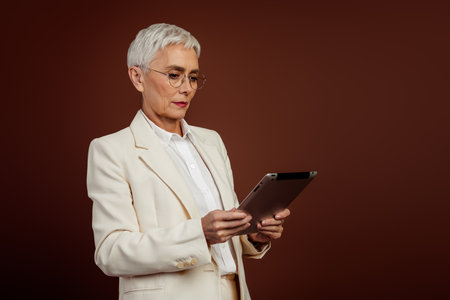 Successful mature businesswoman holding a digital tablet in a formal white suit against a brown background, exuding professionalismの写真素材