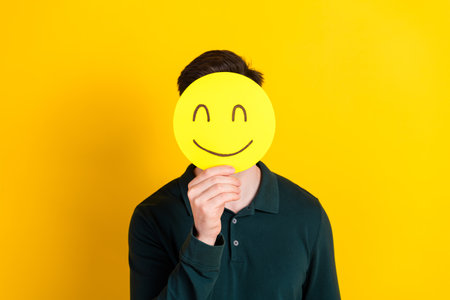 Young guy covering face with smiley on yellow background represents happiness and humorの写真素材