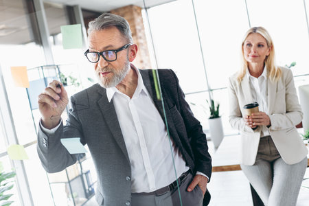 Senior businessman presenting strategies on glass board to colleague in modern office environmentの写真素材