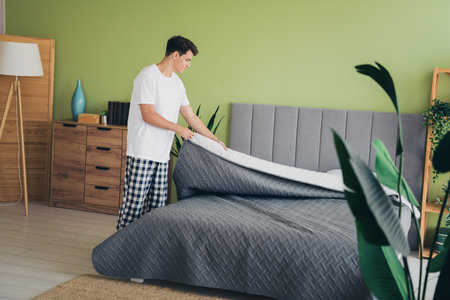 Young male making his bed in a cozy bedroom, preparing for a fresh start to the day in a comfortable...