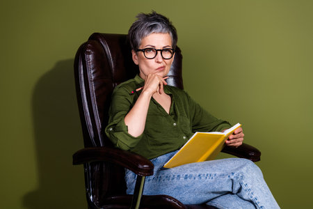 Confident mature woman posing in stylish glasses and green shirt against a khaki background with a book in handの写真素材