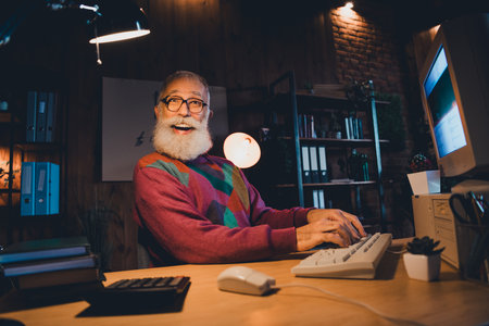 Elderly male programmer working at home office in the evening using a computer, providing a sense of dedication and creativityの写真素材