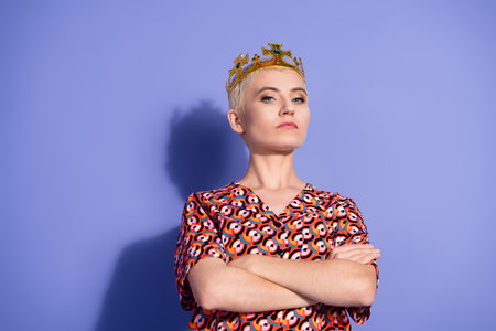 Confident young woman with short blonde hair wearing a geometric print top and a crown on a bright purple backgroundの写真素材