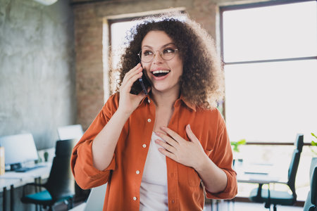 Young smiling businesswoman with curly hair talking on the phone in a bright office setting during daylightの写真素材