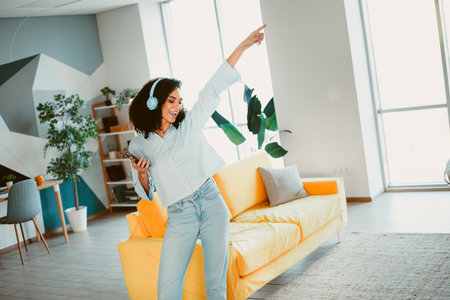 Young cheerful woman enjoying music and dancing in a colorful modern living room with headphones, radiating joy and energy.の写真素材