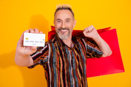 Charismatic mature man with blonde dyed hair showcasing a card while holding a red shopping bag on a...