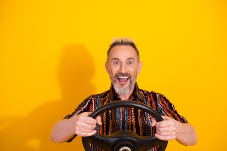 Cheerful mature man with a steering wheel in hand on a bright yellow background showcasing casual style and happinessの写真素材