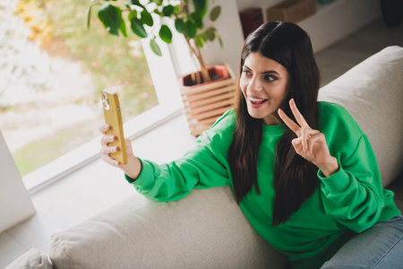 Young woman at home taking a selfie while posing cheerfully in a casual green sweatshirtの写真素材