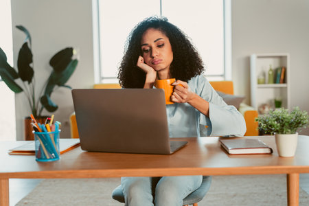 Young woman at home holding a coffee cup and looking at a laptop screen while enjoying a peaceful day in a contemporary and cozy settingの写真素材