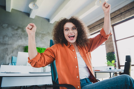Excited young woman celebrating success in a modern workspace with arms raised, showcasing a joyful triumphの写真素材