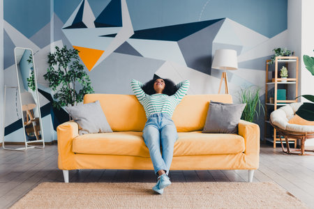 Young woman relaxing on a yellow sofa in a stylish living room with geometric wall art and natural lightingの写真素材