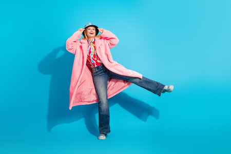 Energetic mature woman in fashionable pink coat smiling and posing dynamically against a vibrant blue studio backgroundの写真素材