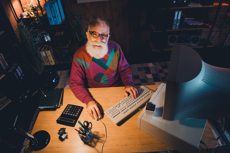 Elderly gentleman working at a desk in a cozy office setting with vintage computer equipment and wooden shelvesの写真素材
