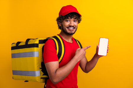 Smiling male delivery service worker in red shirt holding a smartphone with blank screen against vibrant yellow backgroundの写真素材
