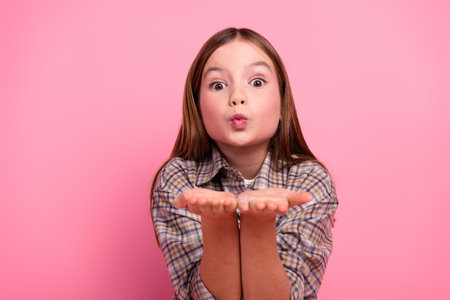 Charming young girl in checkered shirt blowing air kisses against a pink backgroundの写真素材