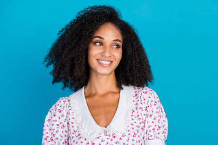 Charming young woman with curly hair in a floral dress smiling against a blue background, capturing beauty and confidence.の写真素材