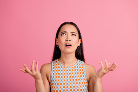 Confused young asian woman looking upwards with puzzled expression on pink background