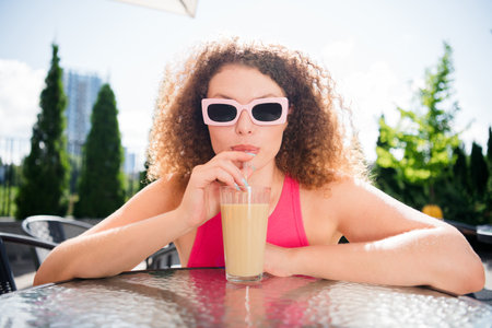 Charming young woman with curly hair and pink sunglasses enjoys a refreshing drink on a sunny day outdoorsの写真素材