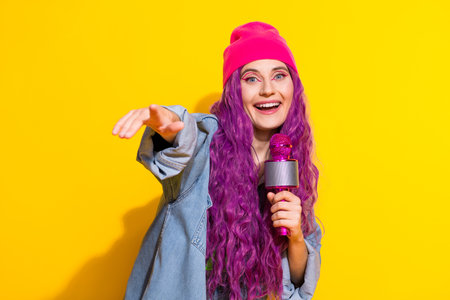 Cheerful young woman with pink hair holding microphone and gesturing against bright yellow background, expressing joy and personalityの写真素材