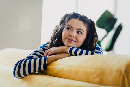 Young woman in striped pullover relaxing on sofa and looking content in bright living room during daylightの写真素材