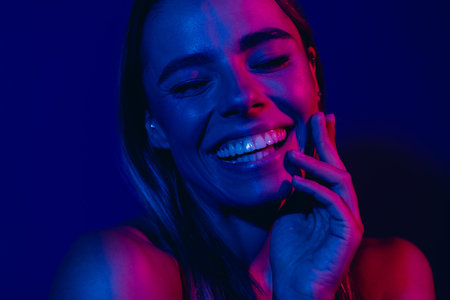 Young woman smiling radiantly under vibrant neon lighting creating a trendy and dynamic ambiance with blue and pink huesの写真素材