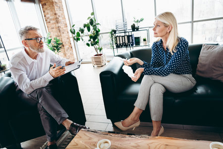 Senior businesspeople engage in a professional conversation in a modern office lounge with natural daylight and stylish interiorの写真素材