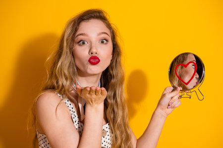 Young Woman Blowing a Kiss With Heart-Shaped Reflection on Mirror Against Vibrant Yellow Backgroundの写真素材