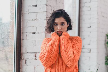 Young woman in an orange sweater standing near a window, exuding cozy and relaxed vibes in a stylish modern home interiorの写真素材