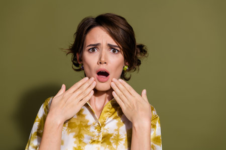 Surprised young woman in elegant blouse with khaki background, expressing shock and disbelief, highlighting modern styleの写真素材