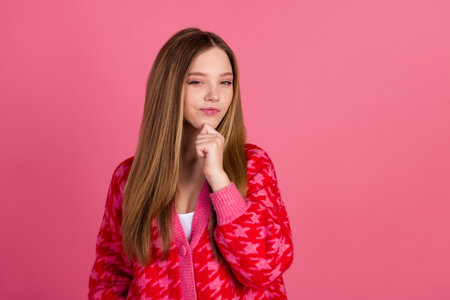 Charming young girl posing with thoughtful expression in casual red cardigan against a pink backgroundの写真素材