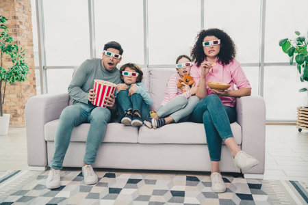 Happy family wearing 3D glasses enjoying a home movie day with snacks in cozy apartment setting on a relaxing weekend afternoon.の写真素材