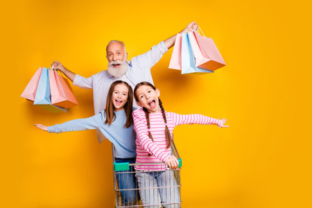 Cheerful grandfather with two happy granddaughters shopping, all smiling brightly, against a vibrant yellow background, celebrating family joy and togethernessの写真素材