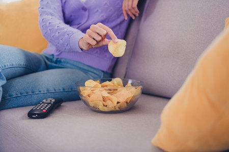 Young woman with blond hair and attractive smile relaxing indoors, enjoying a weekend in a knitted sweater, reaching for chipsの写真素材