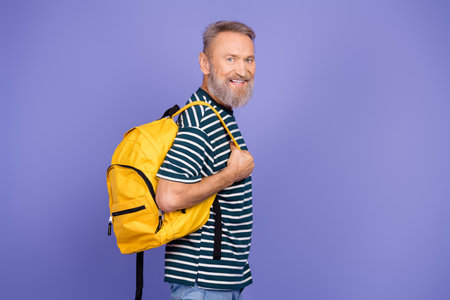 Smiling elderly man with a yellow backpack posing confidently in casual striped shirt against purple background.の写真素材