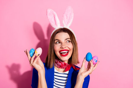 Young woman with bunny ears holding colorful painted eggs against a pink background, celebrating...