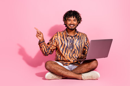 Smiling Young Man Sitting with Laptop on Pink Background Pointing Upwards as a Gesture of Creativity, Leisure, and Multicultural Styleの写真素材